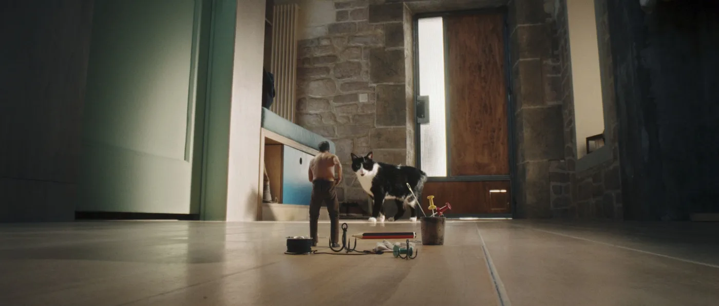 Ein kleiner Mann steht auf einem hellen Fliesenboden vor einer großen Holztür mit Steinmauern. Neben ihm steht eine schwarz-weiße Katze, die deutlich größer als der Mann wirkt. Vor dem Mann liegen verschiedene kleine Gegenstände, darunter ein Eimer und ein Seil.