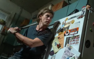 Ein Mann in einem dunkelblauen T-Shirt hält einen Baseballschläger in erhobener Schlagposition. Er steht in einer Küche vor einem Kühlschrank, der mit verschiedenen Aufklebern, Fotos und Zetteln beklebt ist. Im Hintergrund sind Küchenschränke und ein Fenster zu sehen. [erstellt mit KI]