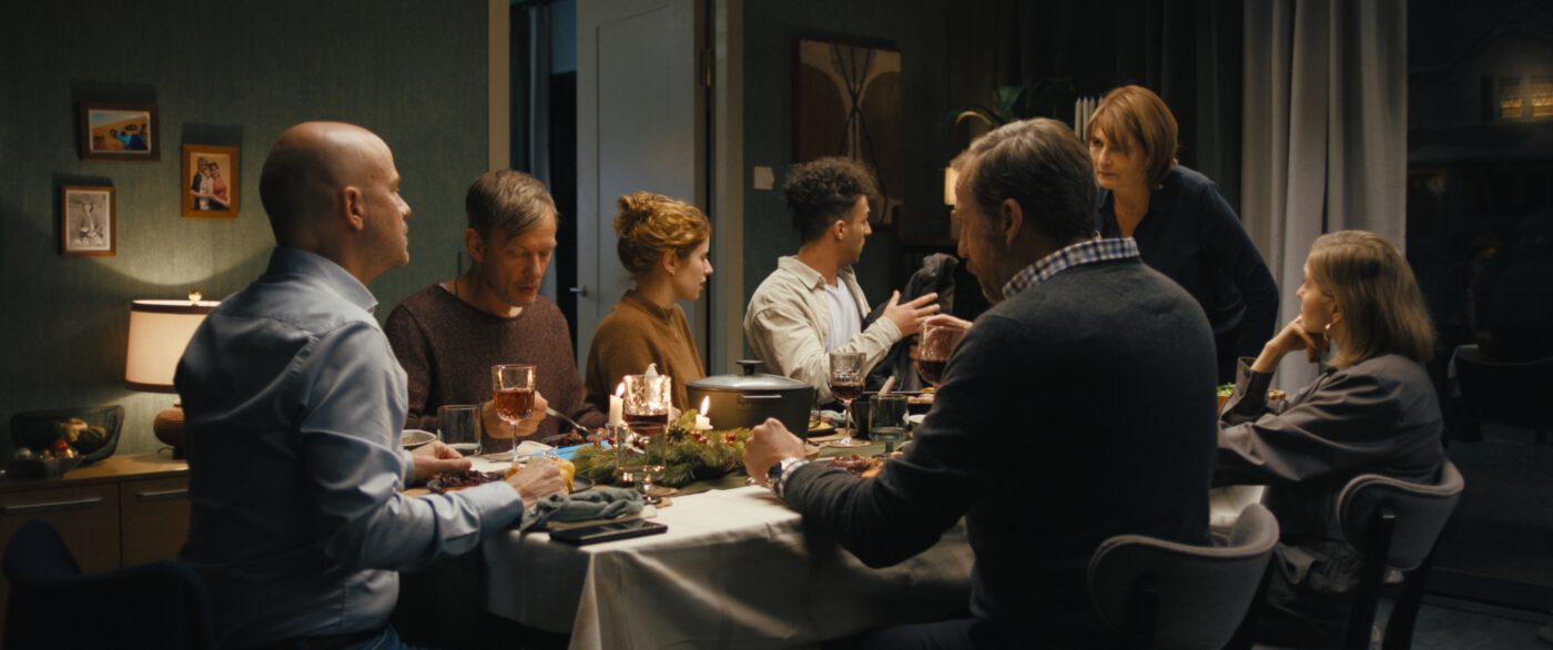 Eine Gruppe von Menschen sitzt an einem gedeckten Esstisch in einem gemütlich eingerichteten Raum. Der Tisch ist mit einem weißen Tischtuch bedeckt und mit Kerzen, Gläsern und Tellern dekoriert. Im Hintergrund sind an der Wand mehrere Bilderrahmen zu sehen. Die Personen sind in Gespräche vertieft und halten teilweise Gläser in der Hand. [erstellt mit KI]