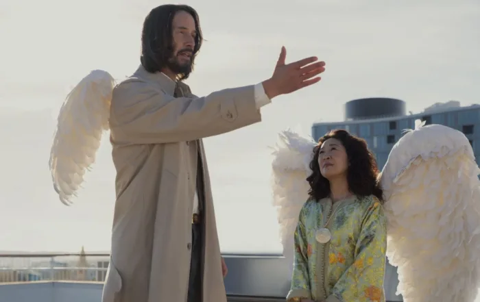 Zwei Personen mit großen weißen Flügeln auf dem Rücken stehen auf einer Dachterrasse. Die linke Person trägt einen hellen Trenchcoat und hebt den rechten Arm mit ausgestreckter Hand. Die rechte Person trägt ein hellgrünes, gemustertes Kleid und hält die Hände vor dem Körper zusammen. Im Hintergrund sind Gebäude und ein bewölkter Himmel zu sehen. [erstellt mit KI]