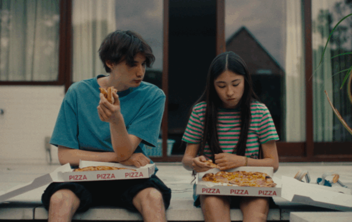 Zwei Personen sitzen nebeneinander auf einer Stufe vor einem Gebäude mit großen Fenstern. Beide halten jeweils ein Stück Pizza in der Hand, vor ihnen stehen geöffnete Pizzakartons mit der Aufschrift 'PIZZA'. Die Person links trägt ein blaues T-Shirt und schwarze Shorts, die Person rechts ein gestreiftes T-Shirt mit kurzen Ärmeln und Shorts. Im Hintergrund ist ein Teil eines Hauses mit einem Giebel zu sehen. [erstellt mit KI]