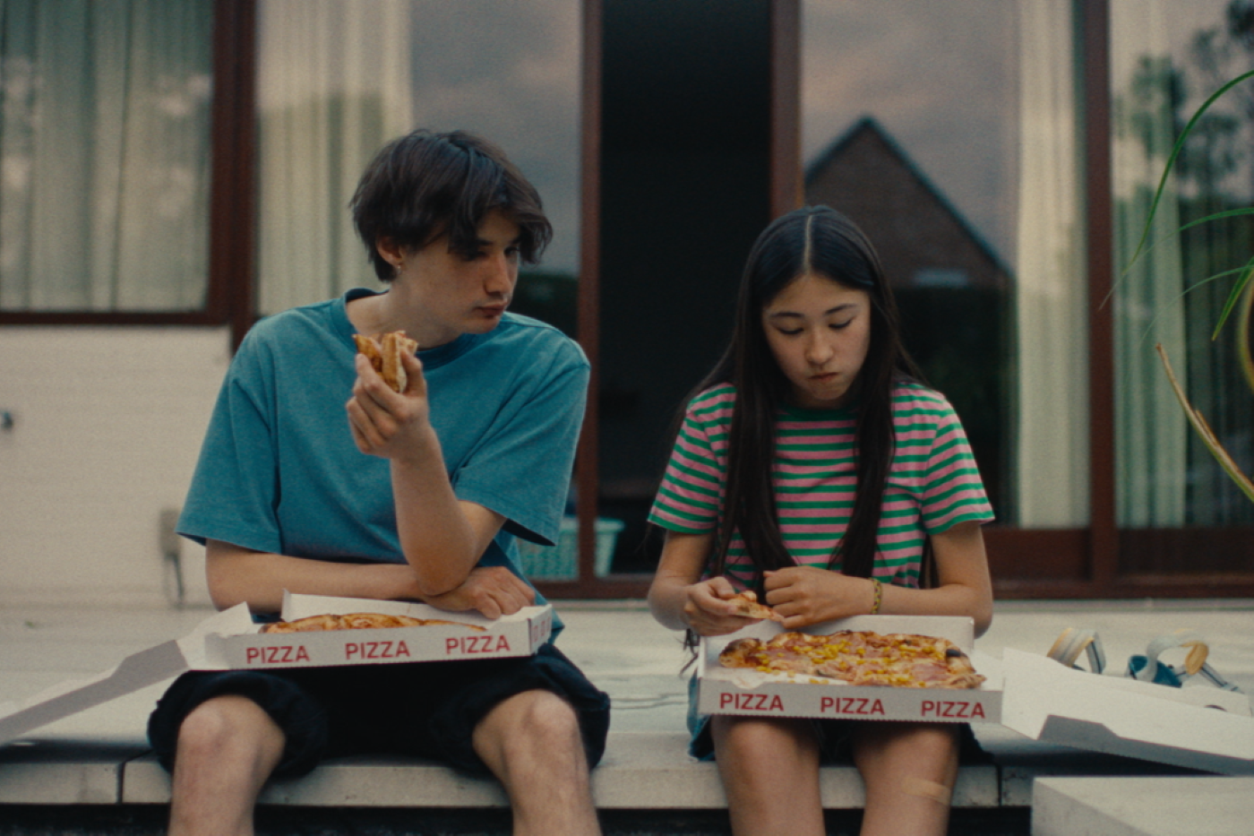 Zwei Personen sitzen nebeneinander auf einer Stufe vor einem Gebäude mit großen Fenstern. Beide halten jeweils ein Stück Pizza in der Hand, vor ihnen stehen geöffnete Pizzakartons mit der Aufschrift 'PIZZA'. Die Person links trägt ein blaues T-Shirt und schwarze Shorts, die Person rechts ein gestreiftes T-Shirt mit kurzen Ärmeln und Shorts. Im Hintergrund ist ein Teil eines Hauses mit einem Giebel zu sehen. [erstellt mit KI]