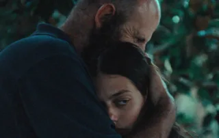 Ein Mann mit dunklem Hemd hält eine junge Frau, seine Tochter, umarmend an sich. Beide sind von Bäumen und Grünpflanzen umgeben. [erstellt mit KI]