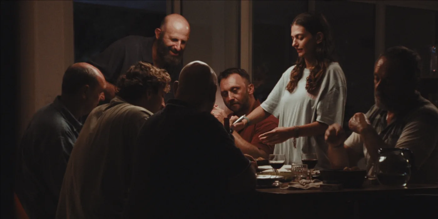 Mehrere Personen sitzen an einem dunklen Holztisch, auf dem Gläser und Teller stehen. Eine junge Frau mit langem, braunem Haar und weißem Oberteil steht am Tisch und reicht etwas an eine sitzende Person weiter. Die Umgebung ist schwach beleuchtet. [erstellt mit KI]