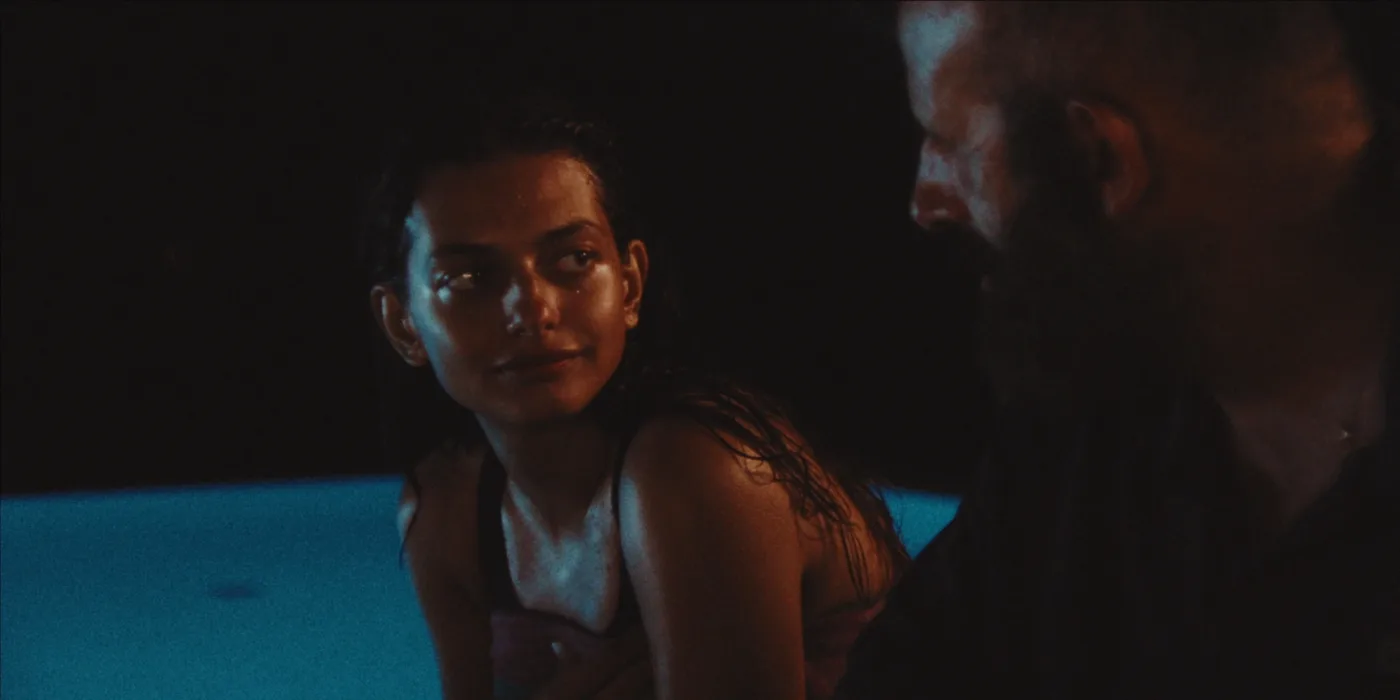 Zwei Personen in dunkler Umgebung, die sich gegenüberstehen. Die Person links trägt ein ärmelloses Oberteil und hat nasse, nach hinten gekämmte Haare. Die Person rechts ist im Profil zu sehen und trägt ein dunkles Hemd. [erstellt mit KI]