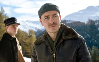 Zwei Männer in Winterkleidung stehen vor einer schneebedeckten Berglandschaft mit Nadelbäumen. Der vordere Mann trägt eine grüne Strickmütze, eine braune Lederjacke mit Fellkragen und einen grünen Pullover. Der hintere Mann trägt eine schwarze Uniformmütze mit rotem Streifen und einen dunklen Mantel. [erstellt mit KI]