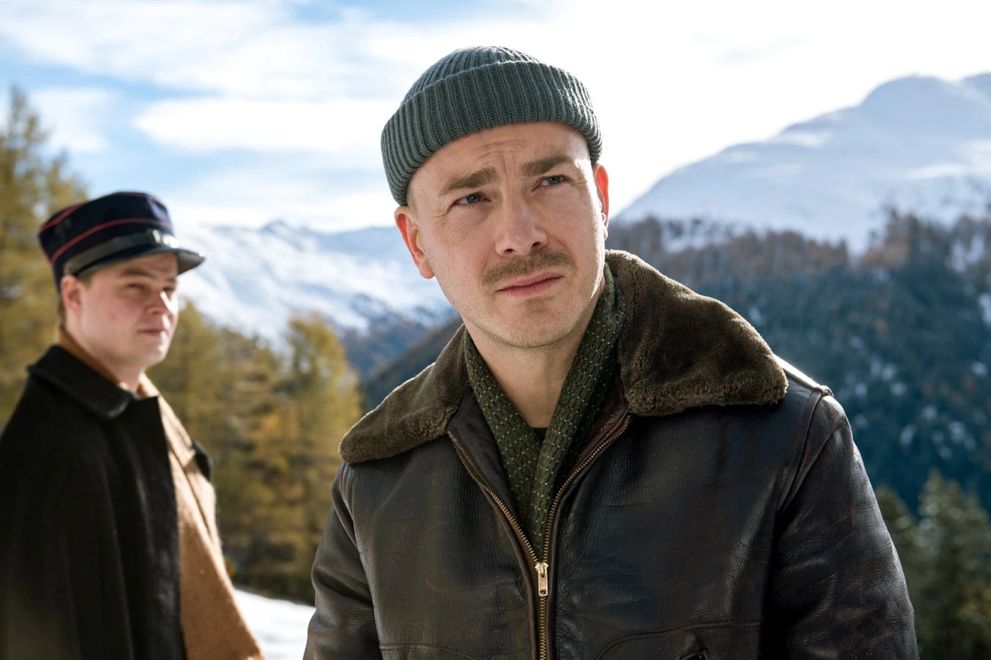 Zwei Männer in Winterkleidung stehen vor einer schneebedeckten Berglandschaft mit Nadelbäumen. Der vordere Mann trägt eine grüne Strickmütze, eine braune Lederjacke mit Fellkragen und einen grünen Pullover. Der hintere Mann trägt eine schwarze Uniformmütze mit rotem Streifen und einen dunklen Mantel. [erstellt mit KI]
