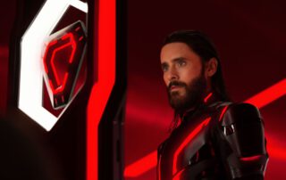 Ein Mann mit dunklem, schulterlangem Haar und Bart steht in einem futuristischen, rot beleuchteten Raum. Er trägt einen schwarzen Anzug mit leuchtenden roten Linien, die an eine Science-Fiction-Rüstung erinnern. Sein Blick ist ernst und fokussiert, er steht vor einem futuristischen Display mit einem roten, leuchtenden Symbol. [erstellt mit KI]