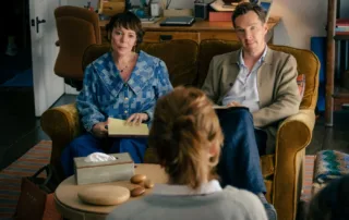 Zwei Personen sitzen auf einem braunen Sofa in einem Wohnzimmer. Die Person links trägt eine blaue Bluse mit Muster und hält ein Notizbuch auf dem Schoß. Die Person rechts trägt ein helles Hemd und eine beige Jacke. Im Vordergrund ist der Rücken einer dritten Person zu sehen, die ein helles Oberteil trägt. Vor ihnen steht ein runder Couchtisch mit einer grauen Taschentuchbox und drei runden Holzobjekten. Im Hintergrund sind ein Schreibtisch mit Laptop und ein Stuhl zu sehen. [erstellt mit KI]