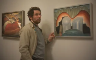 Ein Mann mit lockigem Haar trägt eine beige Jacke über einem grauen Pullover. Er steht vor einer weißen Wand und berührt mit der rechten Hand den Rahmen eines Gemäldes. Das Gemälde zeigt abstrakte Formen in Braun-, Blau- und Grüntönen, darunter zwei große, runde, weiße Objekte. Links an der Wand hängt ein weiteres Gemälde mit einer Darstellung, die runde und gebogene Formen in Rot-, Braun- und Gelbtönen zeigt. [erstellt mit KI]