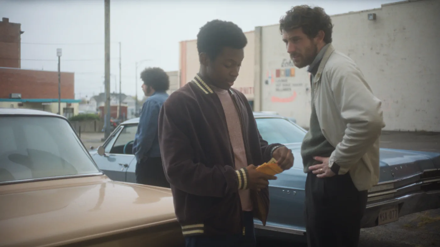 Drei Männer stehen auf einem Parkplatz vor zwei geparkten Autos. Ein Mann trägt eine dunkelblaue Jacke mit gelben Streifen an den Bündchen und einen rosa Pullover. Ein anderer Mann trägt eine helle Jacke und dunkle Hose, die Hände in die Hüften gestützt. Der dritte Mann steht im Hintergrund, trägt eine blaue Jacke und hat lockiges Haar. Im Hintergrund sind Gebäude und ein Zaun zu sehen. [erstellt mit KI]
