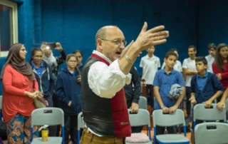 Ein Mann steht im Vordergrund mit erhobenen Händen und dirigiert. Er trägt ein weißes Hemd, eine rote Weste und eine braune Hose. Im Hintergrund stehen mehrere Personen, die auf ihn schauen. Einige sitzen auf Stühlen, andere stehen. Die Wand im Hintergrund ist blau. [erstellt mit KI]