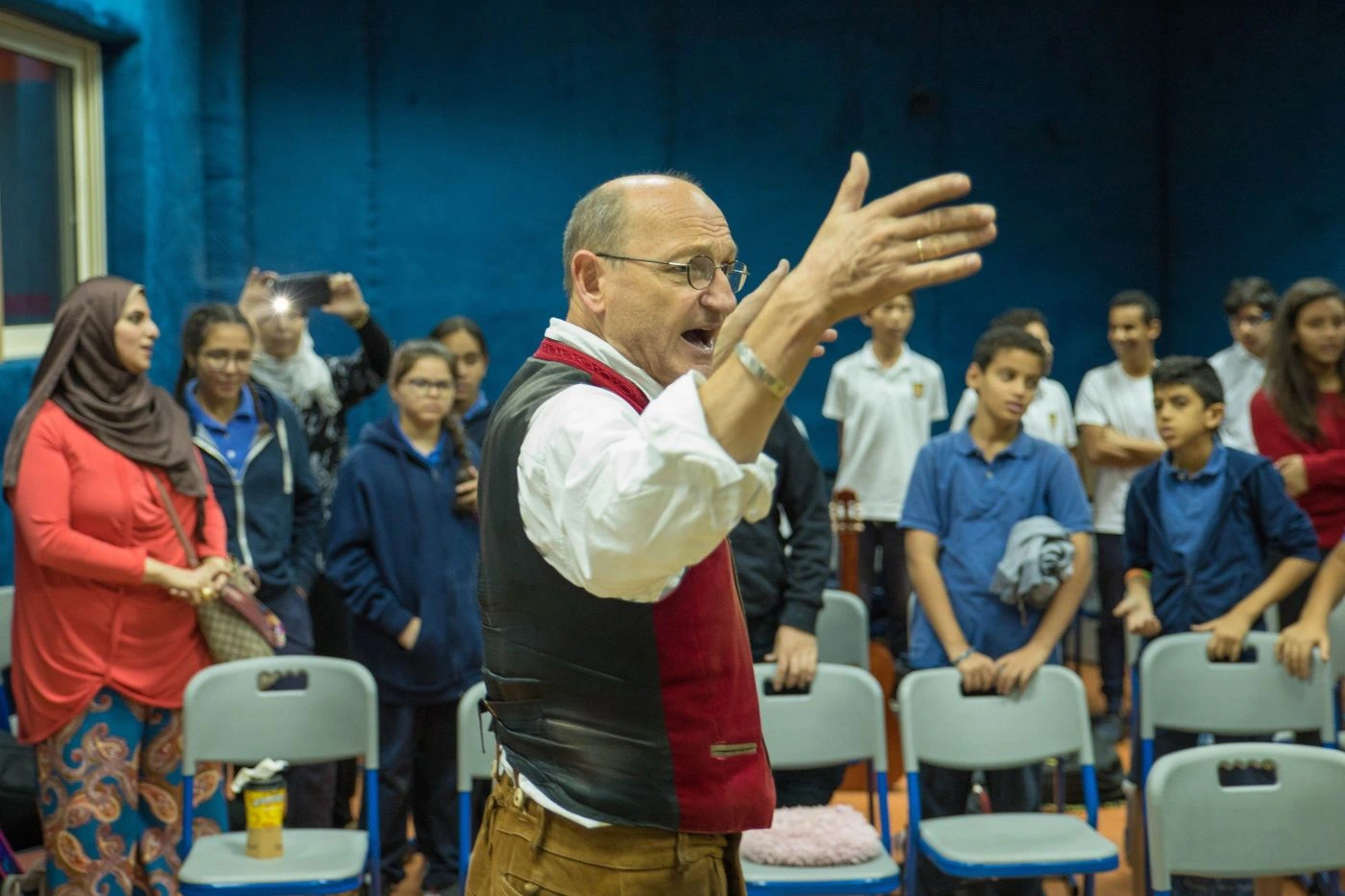 Ein Mann steht im Vordergrund mit erhobenen Händen und dirigiert. Er trägt ein weißes Hemd, eine rote Weste und eine braune Hose. Im Hintergrund stehen mehrere Personen, die auf ihn schauen. Einige sitzen auf Stühlen, andere stehen. Die Wand im Hintergrund ist blau. [erstellt mit KI]