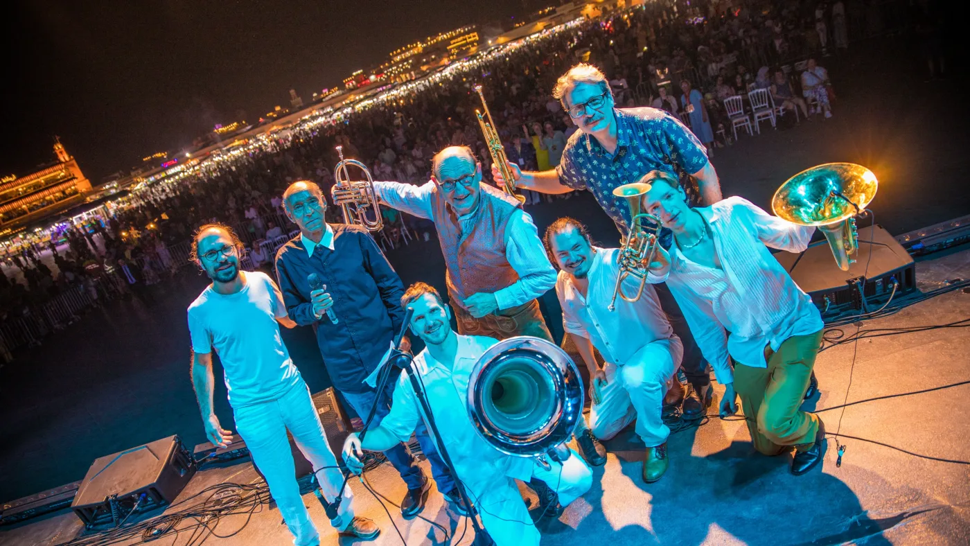 Eine Gruppe von sieben Personen steht auf einer Bühne vor einem großen Publikum bei Nacht. Die Personen halten verschiedene Blechblasinstrumente, darunter Trompeten, Posaunen und eine Tuba. Die Bühne ist mit Lautsprechern und Mikrofonständern ausgestattet. Im Hintergrund sind viele Zuschauer zu sehen, die auf Stühlen sitzen oder stehen. Die Personen tragen legere Kleidung in verschiedenen Farben, darunter Hemden und T-Shirts. [erstellt mit KI]