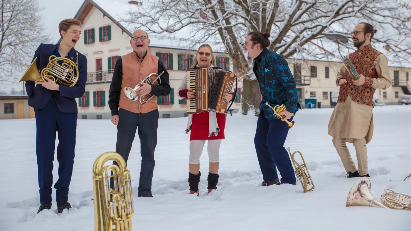 Fünf Personen stehen im Schnee vor einem mehrstöckigen Haus mit grünen Fensterläden. Sie tragen unterschiedliche Kleidung, darunter ein rotes Kleid mit schwarzen Stiefeln, ein blaues Hemd und traditionelle indische Kleidung. Die Personen halten verschiedene Blasinstrumente, darunter eine Trompete, ein Horn, eine Posaune und ein Akkordeon. Im Vordergrund liegt ein großes Blechblasinstrument im Schnee. Im Hintergrund sind schneebedeckte Bäume zu sehen. [erstellt mit KI]