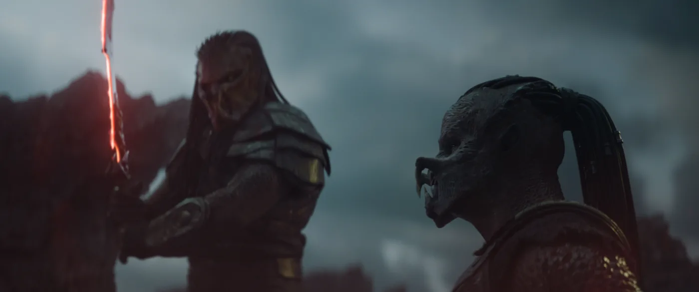 wei humanoide Figuren mit maskenartigen Gesichtern und dunkler, gepanzerter Kleidung stehen vor einem düsteren, bewölkten Himmel. Die vordere Figur trägt eine Maske mit hervorstehenden Kieferzähnen und hat einen Zopf, während die hintere Figur eine Rüstung mit Schulterplatten trägt und ein leuchtendes, rot-orangefarbenes Schwert hält. Im Hintergrund sind unscharfe, dunkle Felsen zu erkennen. [erstellt mit KI]