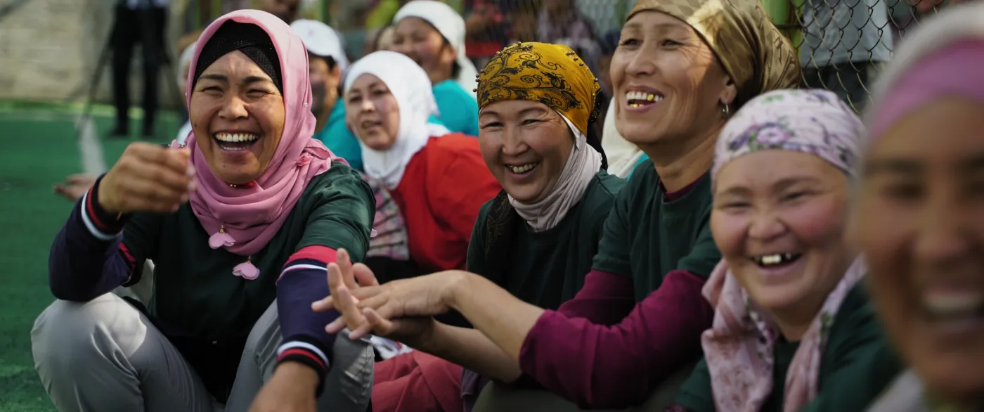 Mehrere Personen sitzen nebeneinander auf dem Boden und lachen. Sie tragen bunte Kopftücher und langärmlige Kleidung. Im Hintergrund ist ein Zaun zu sehen.
