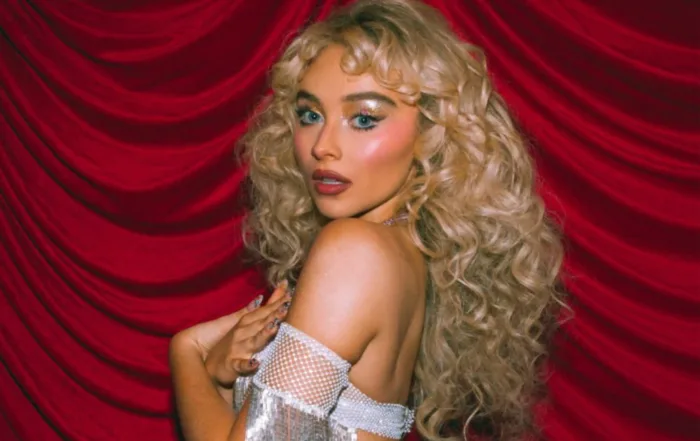 Sabrina Carpenter posiert vor einem dunkelroten, drapierten Vorhang. Sie trägt ein schulterfreies, silbern glitzerndes Outfit mit Fransen und blickt über die Schulter in die Kamera.