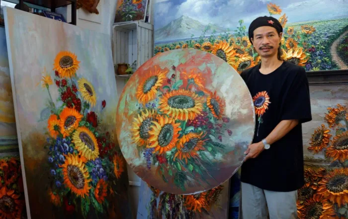 Ein Mann steht in einem Raum voller Sonnenblumen-Gemälde. Er trägt ein schwarzes T-Shirt mit einem Sonnenblumenmotiv und hält ein rundes Gemälde mit Sonnenblumen in den Händen. Im Hintergrund sind weitere Gemälde mit Sonnenblumen zu sehen, darunter ein großes rechteckiges Bild und mehrere kleinere Werke. Die Gemälde zeigen Sonnenblumen in verschiedenen Arrangements und Farben, vor allem in Gelb- und Orangetönen mit grünen Blättern. [erstellt mit KI]
