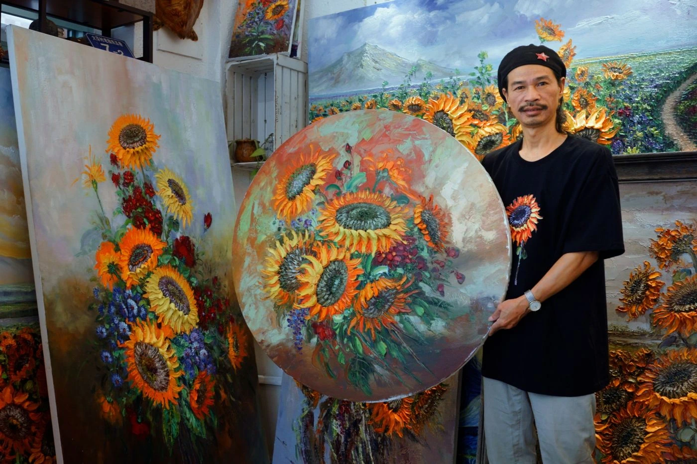 Ein Mann steht in einem Raum voller Sonnenblumen-Gemälde. Er trägt ein schwarzes T-Shirt mit einem Sonnenblumenmotiv und hält ein rundes Gemälde mit Sonnenblumen in den Händen. Im Hintergrund sind weitere Gemälde mit Sonnenblumen zu sehen, darunter ein großes rechteckiges Bild und mehrere kleinere Werke. Die Gemälde zeigen Sonnenblumen in verschiedenen Arrangements und Farben, vor allem in Gelb- und Orangetönen mit grünen Blättern. [erstellt mit KI]