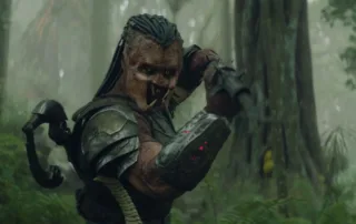 Ein Predator steht in einem nebligen Wald. Er trägt eine schwere, metallische Rüstung mit Schulterpanzer und einen gepanzerten Unterarm, auf dem drei rote Lichter leuchten. Sein rechter Arm ist angewinkelt, die Hand befindet sich nahe am Kopf. Der Hintergrund zeigt dichte Bäume und grünes Unterholz in einer feuchten, nebligen Atmosphäre. [erstellt mit KI]