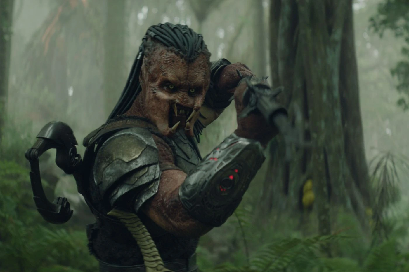 Ein Predator steht in einem nebligen Wald. Er trägt eine schwere, metallische Rüstung mit Schulterpanzer und einen gepanzerten Unterarm, auf dem drei rote Lichter leuchten. Sein rechter Arm ist angewinkelt, die Hand befindet sich nahe am Kopf. Der Hintergrund zeigt dichte Bäume und grünes Unterholz in einer feuchten, nebligen Atmosphäre. [erstellt mit KI]