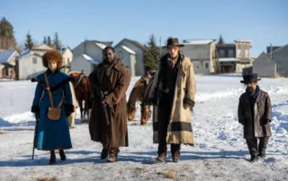 Vier Personen gehen auf einem schneebedeckten Feld, im Hintergrund sind mehrere kleine Häuser und Nadelbäume zu sehen. Die erste Person trägt einen langen blauen Mantel mit schwarzem Pelzbesatz und eine braune Umhängetasche. Die zweite Person ist in einen langen braunen Mantel gehüllt und hält einen Stock. Die dritte Person trägt einen langen, hellen Mantel mit dunklem Besatz und einen Hut. Die vierte Person, ein Kind, trägt einen dunklen Mantel und einen Zylinderhut. Im Hintergrund sind zwei Pferde zu erkennen. [erstellt mit KI]