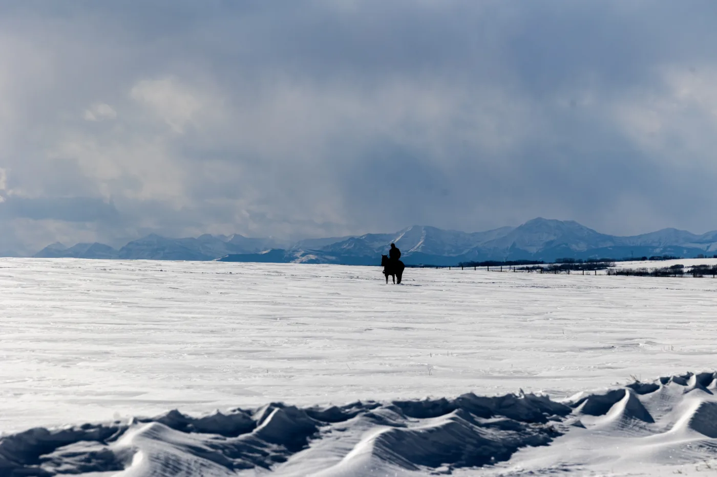 Eine weite, schneebedeckte Landschaft mit einer einzelnen Person auf einem Pferd in der Ferne. Der Himmel ist bewölkt und die Umrisse von Bergen sind am Horizont sichtbar. [erstellt mit KI]