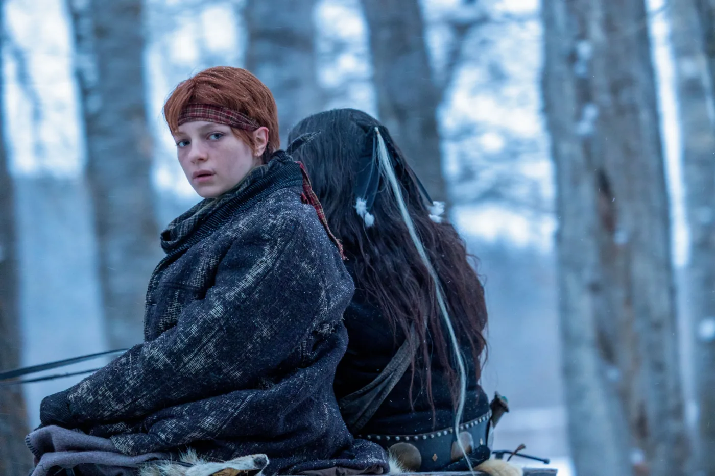 Zwei Personen sitzen Rücken an Rücken in einem Wald mit kahlen Bäumen. Die Person links trägt einen dunklen, grob gewebten Mantel und hat rotbraunes Haar. Die Person rechts hat langes, dunkles Haar mit einigen hellen Strähnen und trägt einen schwarzen Gürtel mit Metallnieten. Beide sitzen auf einem mit Fell bedeckten Untergrund.