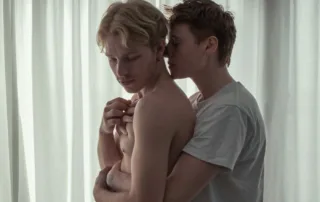 Zwei junge Männer stehen eng umschlungen vor einem hellen, halbtransparenten Vorhang. Der vordere Mann ist oberkörperfrei, der hintere trägt ein weißes T-Shirt. Die Arme des hinteren Mannes umschließen den vorderen, dessen Hände die des anderen halten. [erstellt mit KI]