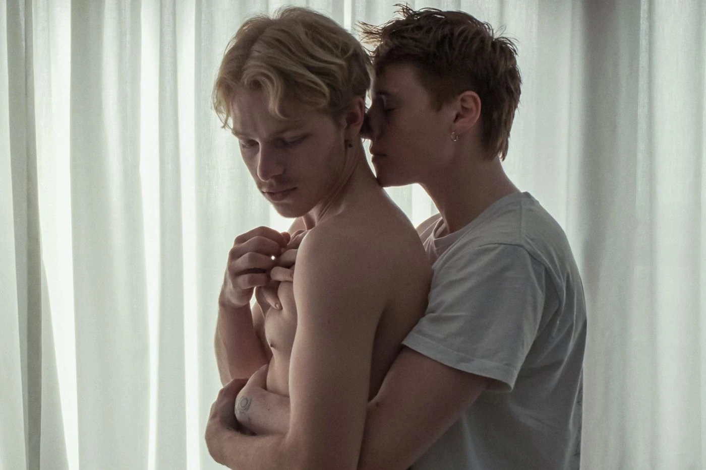 Zwei junge Männer stehen eng umschlungen vor einem hellen, halbtransparenten Vorhang. Der vordere Mann ist oberkörperfrei, der hintere trägt ein weißes T-Shirt. Die Arme des hinteren Mannes umschließen den vorderen, dessen Hände die des anderen halten. [erstellt mit KI]