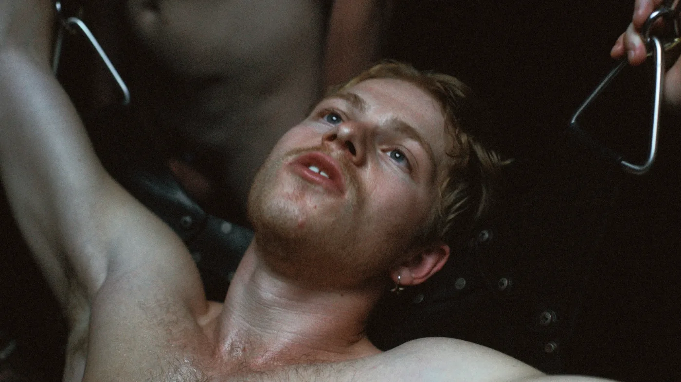 Ein Mann liegt auf dem Rücken auf einer schwarzen Liege oder Bank. Er trägt keine Oberbekleidung und hat die Arme seitlich ausgestreckt. In jeder Hand hält er ein Metallgriffstück, das an Seilen oder Kabeln befestigt ist. Der Hintergrund ist dunkel. [erstellt mit KI]