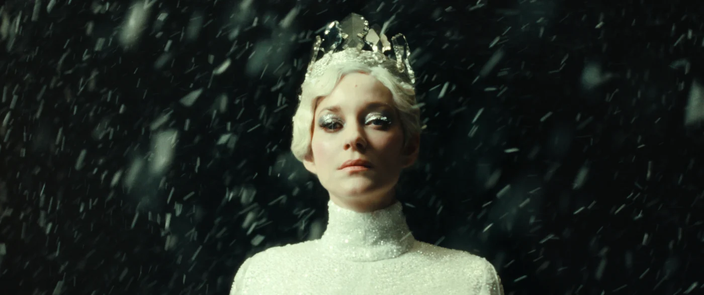 Eine Person trägt ein glitzerndes, weißes, hochgeschlossenes Oberteil. Auf dem Kopf befindet sich eine Eiskrone oder eine skulpturale Kopfbedeckung, die an gefrorenes Wasser erinnert. Im Hintergrund und im Vordergrund sind zahlreiche Schneeflocken oder Eiskristalle zu sehen, die durch die Luft wirbeln. Der Hintergrund ist dunkel gehalten, wodurch die weißen Elemente besonders hervortreten. [erstellt mit KI]