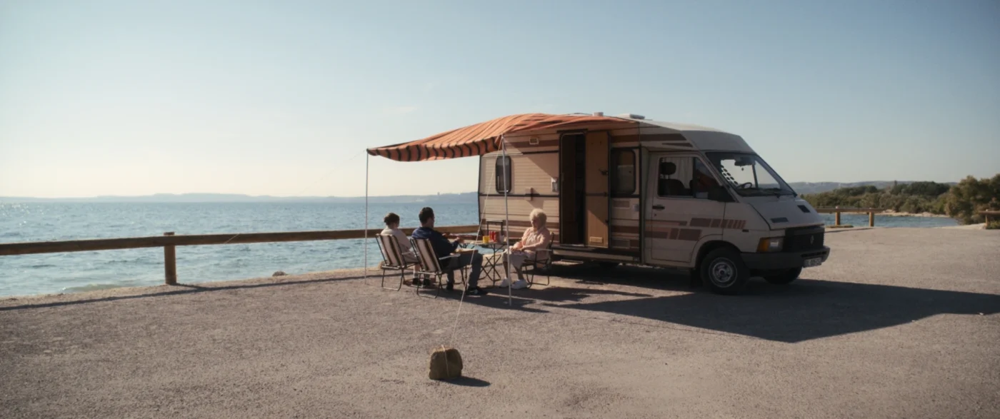 Ein Campingbus mit ausgefahrener Markise steht auf einem Parkplatz direkt am Wasser. Unter der Markise sitzen mehrere Personen auf Campingstühlen um einen kleinen Tisch. Im Hintergrund ist eine ruhige Wasserfläche und ein klarer Himmel zu sehen. [erstellt mit KI]
