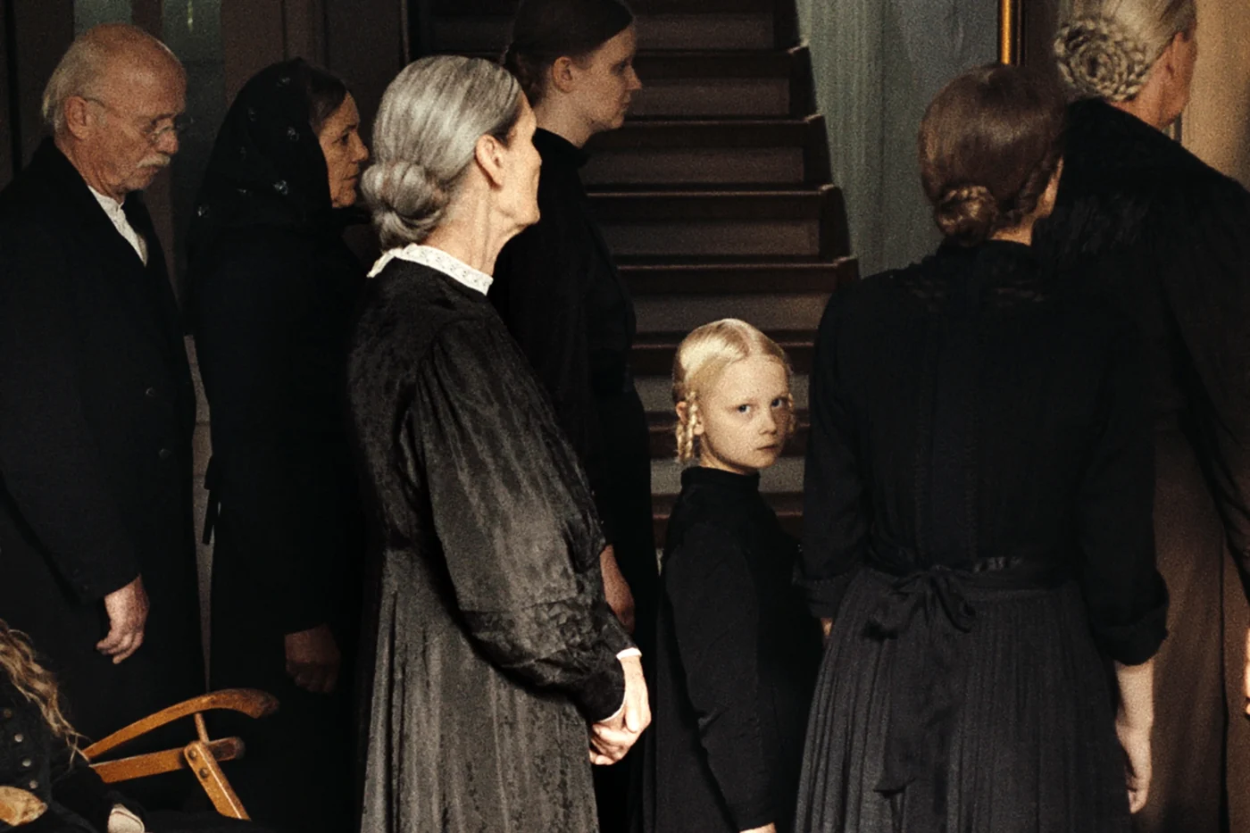 Mehrere Personen in dunkler, altmodischer Kleidung stehen in einem Innenraum vor einer Treppe. Eine Frau mit grauem, streng zurückgebundenem Haar steht seitlich und trägt ein langes schwarzes Kleid. Ein Kind mit blondem Haar steht in der Mitte. Die Szene wirkt ruhig und ernst, die Beleuchtung ist gedämpft. [erstellt mit KI]
