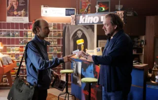 Zwei Männer stehen sich in einem Raum gegenüber, der wie ein Laden oder eine kleine Bibliothek wirkt. Der Mann links trägt eine blaue Jacke und eine Umhängetasche, er hält eine DVD-Hülle in der Hand und reicht sie dem Mann rechts, der einen dunkelblauen Pullover trägt. Im Hintergrund sind Regale mit weiteren DVDs oder Büchern sowie verschiedene Plakate und Schilder zu sehen, darunter ein Schild mit der Aufschrift "kino". Der Raum ist gemütlich eingerichtet mit einem Teppich und zwei grünen Hockern. [erstellt mit KI]