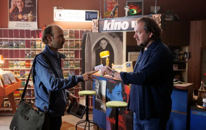 Zwei Männer stehen sich in einem Raum gegenüber, der wie ein Laden oder eine kleine Bibliothek wirkt. Der Mann links trägt eine blaue Jacke und eine Umhängetasche, er hält eine DVD-Hülle in der Hand und reicht sie dem Mann rechts, der einen dunkelblauen Pullover trägt. Im Hintergrund sind Regale mit weiteren DVDs oder Büchern sowie verschiedene Plakate und Schilder zu sehen, darunter ein Schild mit der Aufschrift "kino". Der Raum ist gemütlich eingerichtet mit einem Teppich und zwei grünen Hockern. [erstellt mit KI]