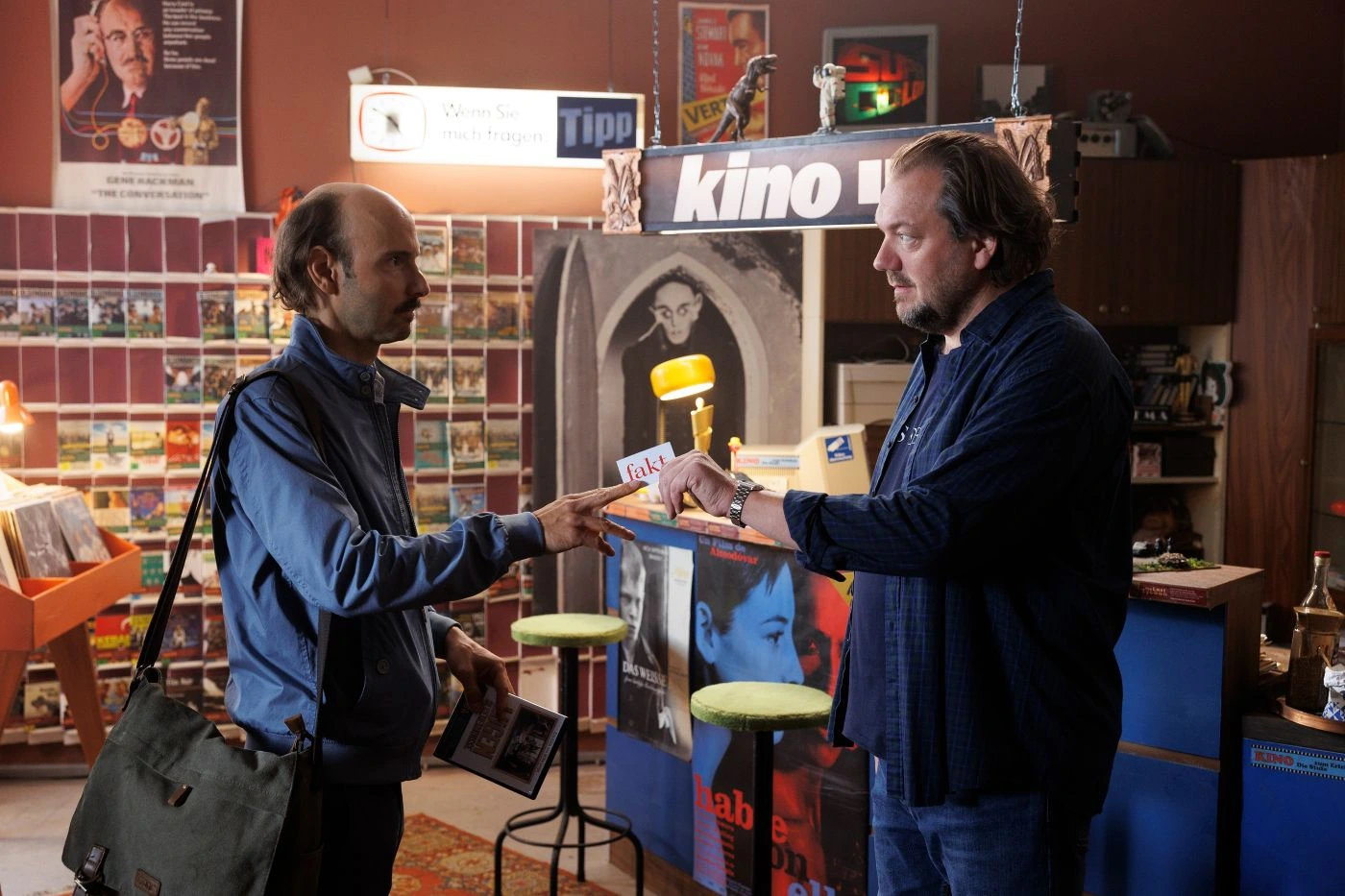 Zwei Männer stehen sich in einem Raum gegenüber, der wie ein Laden oder eine kleine Bibliothek wirkt. Der Mann links trägt eine blaue Jacke und eine Umhängetasche, er hält eine DVD-Hülle in der Hand und reicht sie dem Mann rechts, der einen dunkelblauen Pullover trägt. Im Hintergrund sind Regale mit weiteren DVDs oder Büchern sowie verschiedene Plakate und Schilder zu sehen, darunter ein Schild mit der Aufschrift "kino". Der Raum ist gemütlich eingerichtet mit einem Teppich und zwei grünen Hockern. [erstellt mit KI]