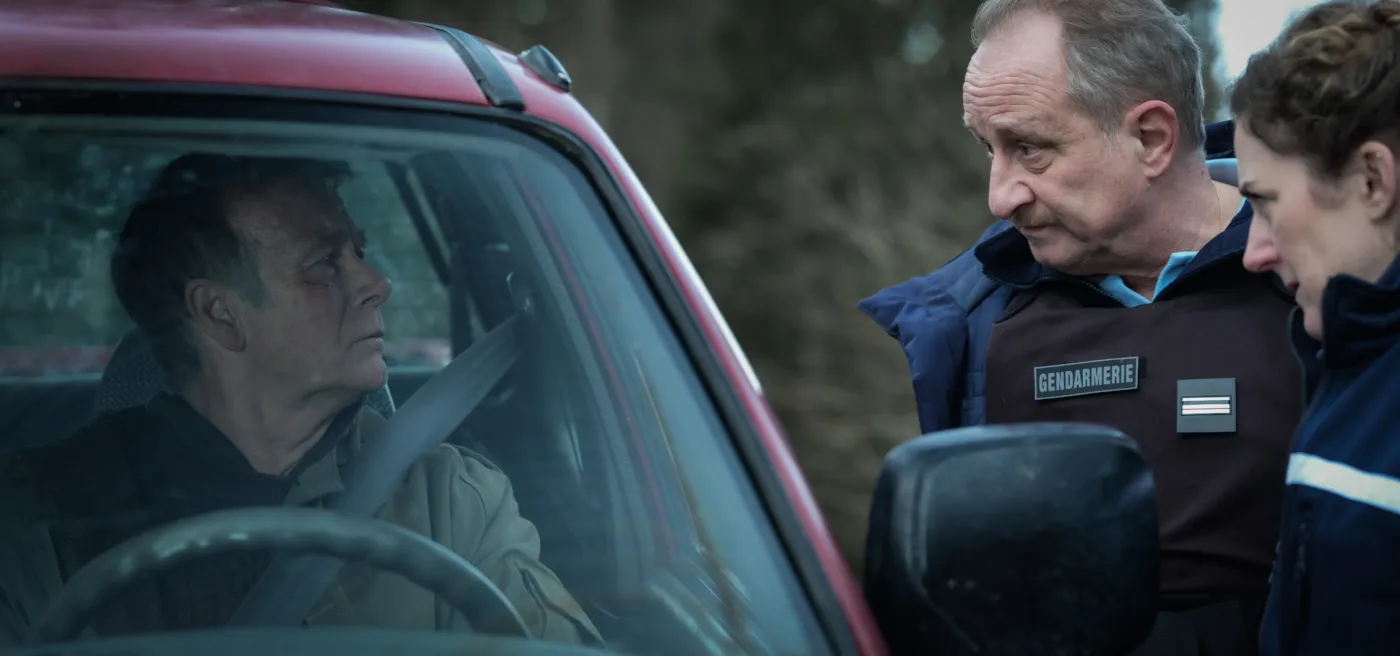 Ein Mann sitzt angeschnallt auf dem Fahrersitz eines roten Autos. Vor dem Auto steht ein Polizist in blauer Uniform mit der Aufschrift "GENDARMERIE" auf der Weste. Der Polizist spricht mit dem Mann im Auto. Im Hintergrund sind unscharfe Bäume zu erkennen. [erstellt mit KI]