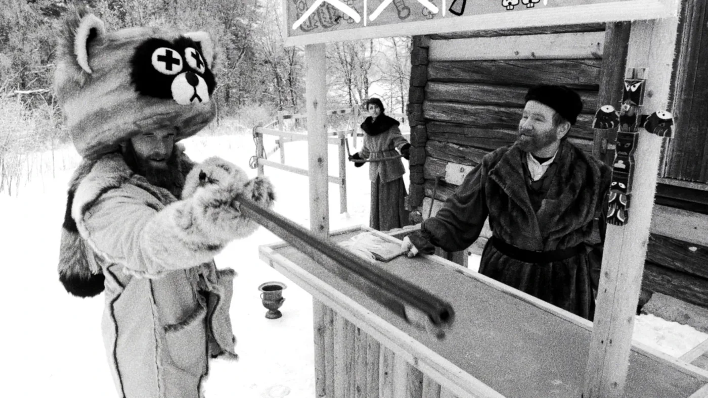 Szene in schwarz-weiß mit drei Personen in dicken, pelzbesetzten Mänteln vor einer Holzhütte im Schnee. Links trägt eine Person ein auffälliges Kostüm mit einer großen Tiermütze, die ein stilisiertes Bibergesicht mit gekreuzten Augen zeigt. Diese Person hält einen langen Stock oder eine Stange über einen Tresen, hinter dem eine weitere Person steht. Im Hintergrund steht eine dritte Person mit einem Gegenstand in der Hand. Über dem Tresen hängt ein Schild mit Symbolen. [erstellt mit KI]