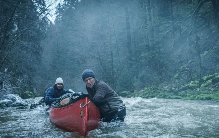 Zwei Personen stehen in einem reißenden Fluss, der von dichtem Wald umgeben ist. Sie halten ein rotes Kanu, das sie offenbar durch das Wasser manövrieren. Beide tragen wetterfeste Kleidung und Mützen, die Umgebung wirkt neblig und kühl. [erstellt mit KI]