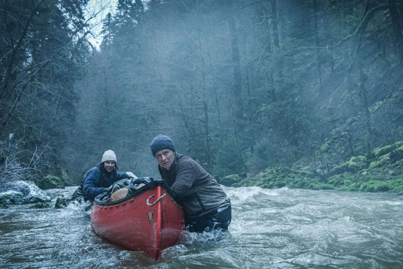 Zwei Personen stehen in einem reißenden Fluss, der von dichtem Wald umgeben ist. Sie halten ein rotes Kanu, das sie offenbar durch das Wasser manövrieren. Beide tragen wetterfeste Kleidung und Mützen, die Umgebung wirkt neblig und kühl. [erstellt mit KI]