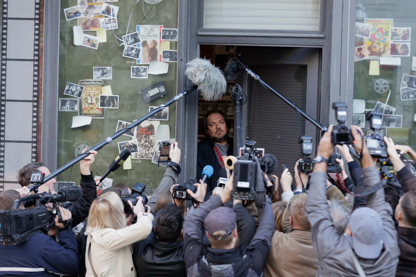 Vor einem Gebäude mit grüner Fassade steht eine Person in einem offenen Türrahmen. Vor der Person versammeln sich zahlreiche Fotografen und Kameraleute mit Kameras, Mikrofonen und Blitzgeräten, die auf die Person gerichtet sind. An der Fassade sind verschiedene Fotos und Filmstreifen als Dekoration angebracht. [erstellt mit KI]
