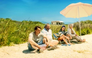 Vier Personen sitzen nebeneinander auf Campingstühlen am Strand. Im Hintergrund stehen hohe Gräser und ein Wohnmobil. Ein großer Sonnenschirm spendet Schatten. Der Himmel ist wolkenlos und blau. [erstellt mit KI]