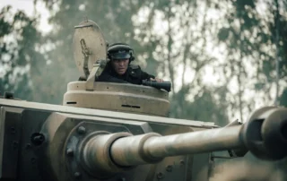 Ein Panzer mit geöffnetem Turmluk steht im Vordergrund. Ein Mensch trägt einen dunklen Helm und sitzt im Turm. Im Hintergrund sind unscharfe Bäume zu erkennen. Die Szene wirkt leicht neblig oder dunstig. [erstellt mit KI]