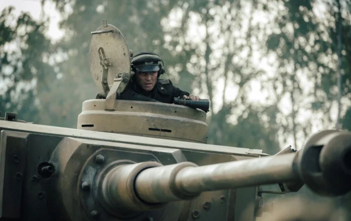 Ein Panzer mit geöffnetem Turmluk steht im Vordergrund. Ein Mensch trägt einen dunklen Helm und sitzt im Turm. Im Hintergrund sind unscharfe Bäume zu erkennen. Die Szene wirkt leicht neblig oder dunstig. [erstellt mit KI]