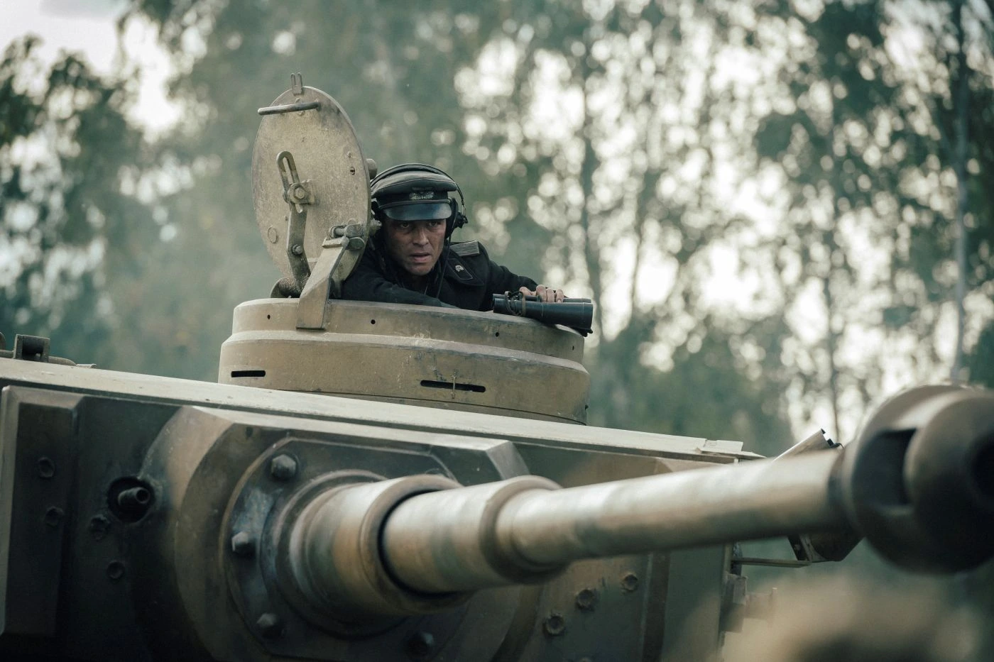 Ein Panzer mit geöffnetem Turmluk steht im Vordergrund. Ein Mensch trägt einen dunklen Helm und sitzt im Turm. Im Hintergrund sind unscharfe Bäume zu erkennen. Die Szene wirkt leicht neblig oder dunstig. [erstellt mit KI]