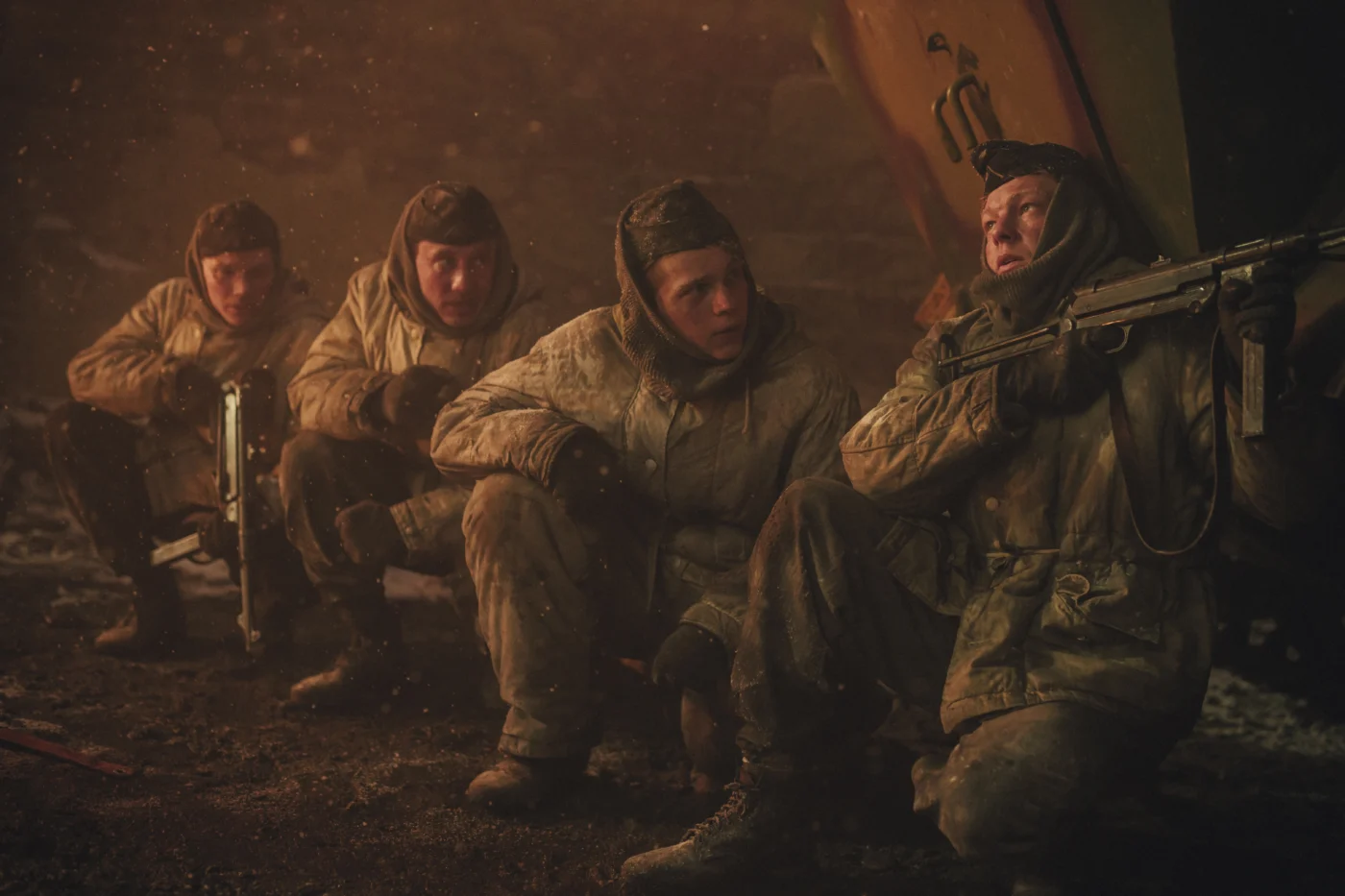 Vier Personen in dicken, hellen Wintermänteln hocken nebeneinander auf dem Boden. Zwei von ihnen halten Gewehre in den Händen. Im Hintergrund sind dunkle, unscharfe Strukturen und ein orangefarbenes Objekt zu erkennen. Die Szene ist in warmes, rötliches Licht getaucht. [erstellt mit KI]