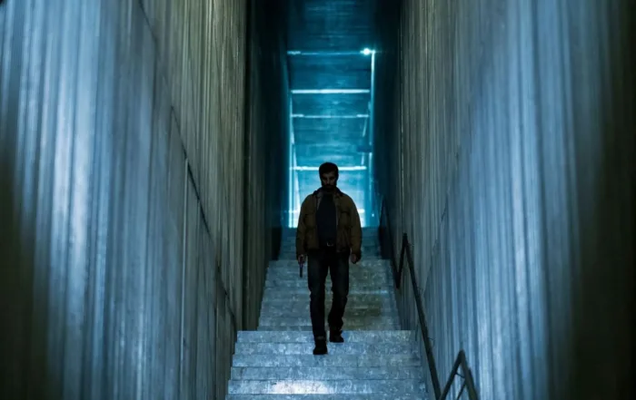 Eine Person mit dunkler Jacke und Hose geht eine breite Treppe in einem schmalen, hohen Gang hinunter. Die Wände und Stufen bestehen aus Beton, das Licht ist kühl und bläulich. Am oberen Ende der Treppe ist ein heller, rechteckiger Lichtschein sichtbar. [erstellt mit KI]