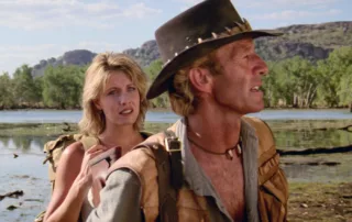 Zwei Personen stehen in einer natürlichen Landschaft mit Wasser und Bäumen im Hintergrund. Die vordere Person trägt einen braunen Hut mit Nieten, ein hellblaues Hemd und eine braune Weste. Um den Hals trägt sie eine Kette mit einem kleinen Anhänger. Die hintere Person hat blonde Haare und trägt einen Rucksack. Im Hintergrund sind Felsen und bewaldete Hügel zu sehen. [erstellt mit KI]